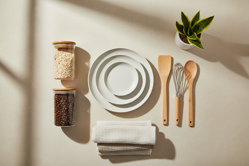 Flat lay of modern home and kitchen essentials including ceramic plates, wooden utensils, glass jars, neutral-toned kitchen towels, and a small indoor plant, arranged neatly on a light beige background, minimal composition, soft natural daylight, subtle shadows, clean aesthetic, high-resolution eCommerce photography, no text, no people, no logos